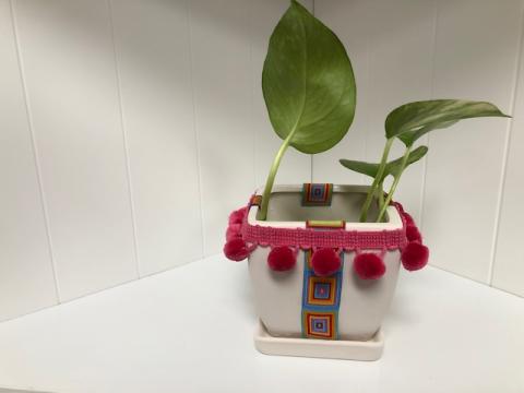 A color photo of a flower pot decorated with patterned tape and pom-pom trim. A small plant is in the pot.