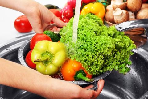 A color photo of a colander of peppers, tomatoes and lettuce being held under a running faucet.