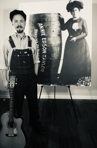 A sepia-toned photo of John John Brown and his guitar. He is is period dress and is standing in front of a photo of a woman with a barrel.