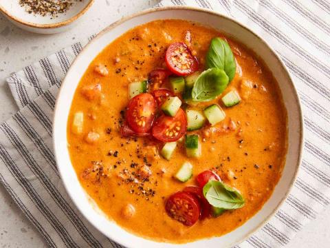 A color photo of a white bowl filled with gazpacho soup that is garnished with diced cucumber and halved cherry tomatoes.