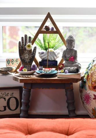 A color photo of a special space in a home, featuring a table with statues, plants and shallow bowls with flowers and an orange cushion in the foreground.