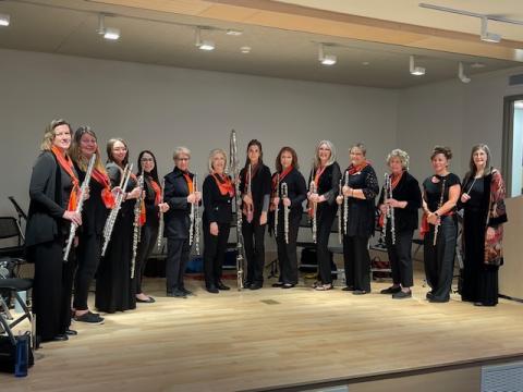A photo of the members of the Long Island Flute Club Choir holding their flutes and standing in a semi-circle on a stage.