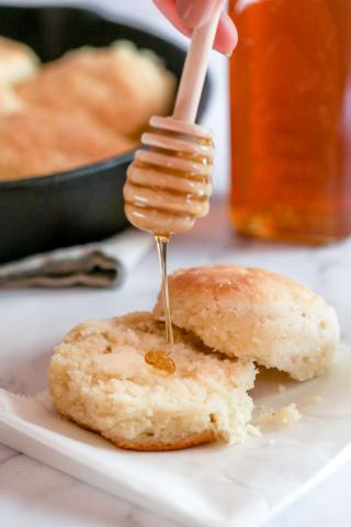 A photo of a wooden honey dipper drizzling honey on top of a cut biscuit.