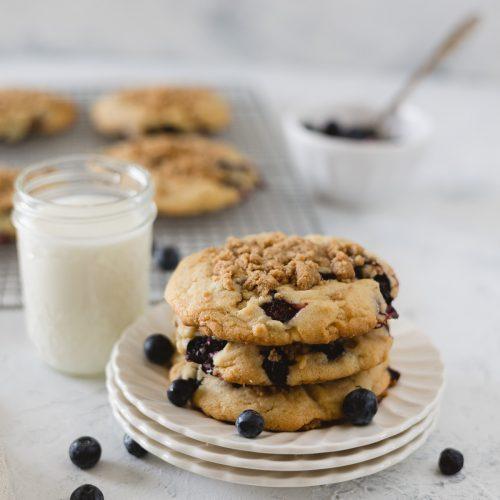 A color photo of blueberry cookies on a white plate accompanied by a glass of milk.