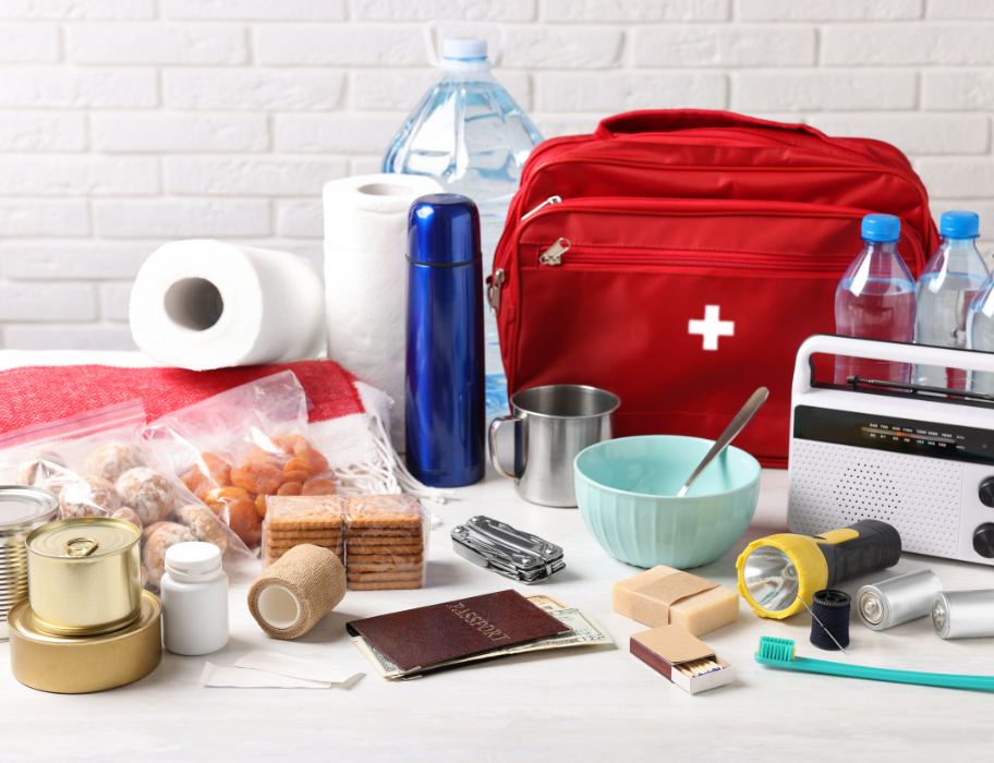  A collection of emergency preparedness items is laid out on a table, including a red first-aid kit, canned goods, water bottles, a flashlight, and a portable radio. The items are arranged against a white brick wall, suggesting a stockpile for a disaster or survival situation.
