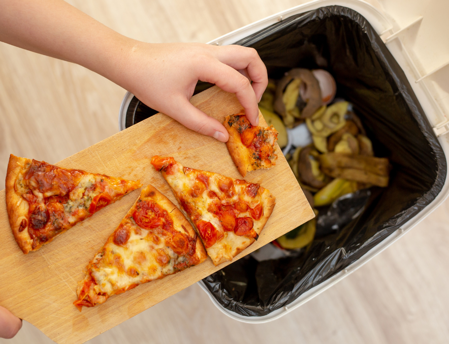 A hand holding a wooden cutting board with leftover slices of pizza drops a crust into a kitchen garbage can lined with a black bag and containing compostable food scraps.
