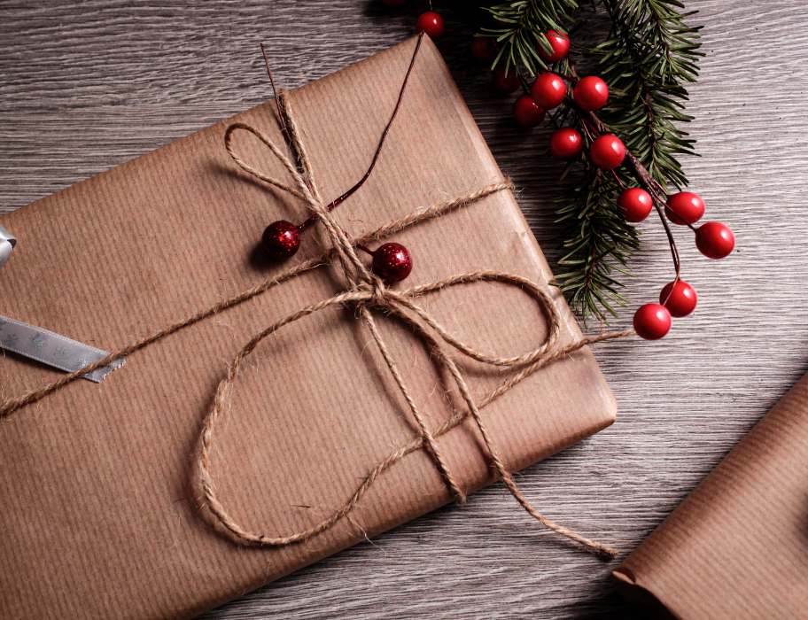  A close-up of a Christmas present wrapped in plain brown craft paper and tied with natural twine. Two small, red glittery berries are attached to the bow. In the upper right corner, there is a sprig of evergreen with bright red faux berries. The gift is resting on a wooden surface.